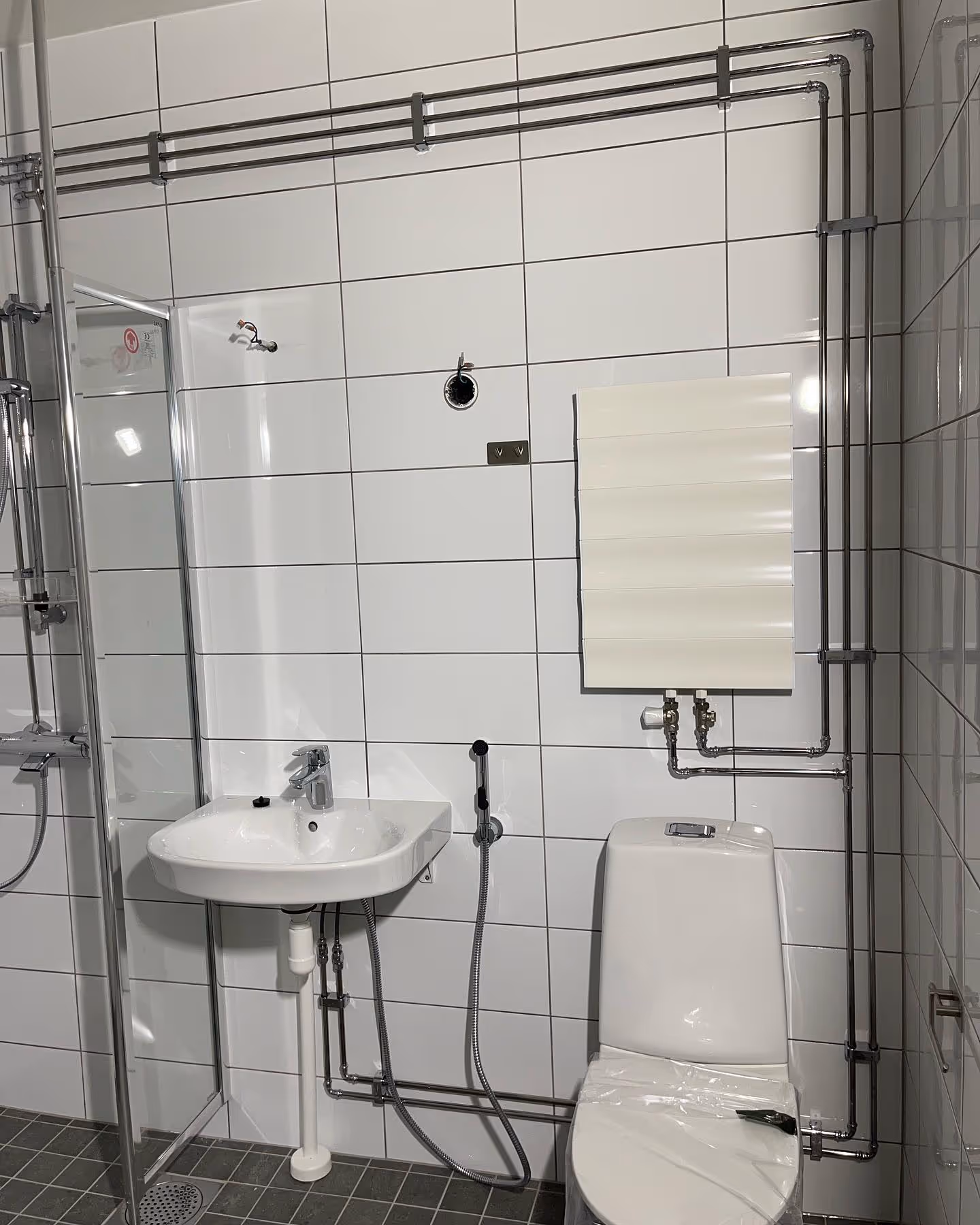 Kylpyhuone, jossa on valkoiset kaakeliseinät, suihku, pesuallas, wc-istuin ja näkyvät LVI-putket.