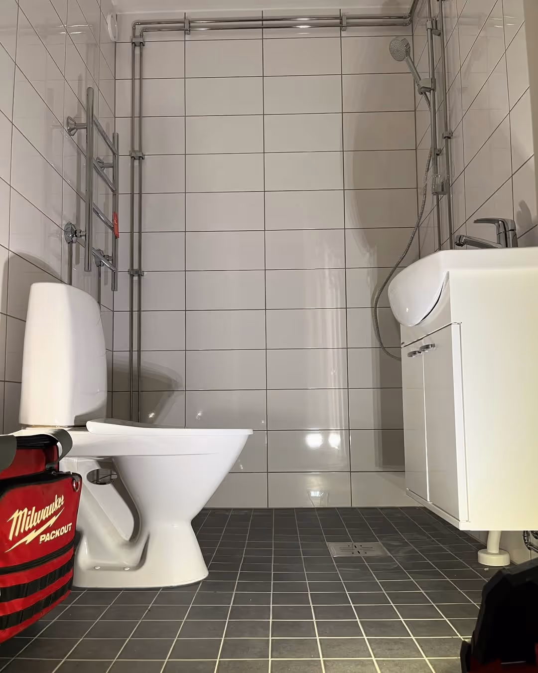 Kylpyhuone, jossa on valkoiset seinälaatat, tummat lattialaatat, wc-istuin, pesuallas ja näkyvillä olevat putkistot.