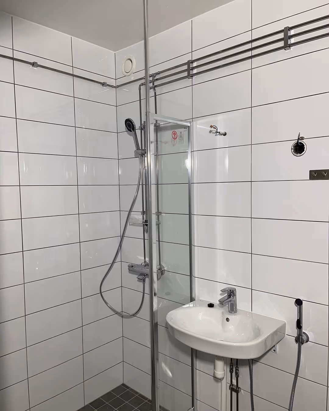 Kylpyhuone, jossa on valkoiset kaakeliseinät, suihku lasiseinällä, seinäkiinnitteinen pesuallas ja näkyvä putkisto.