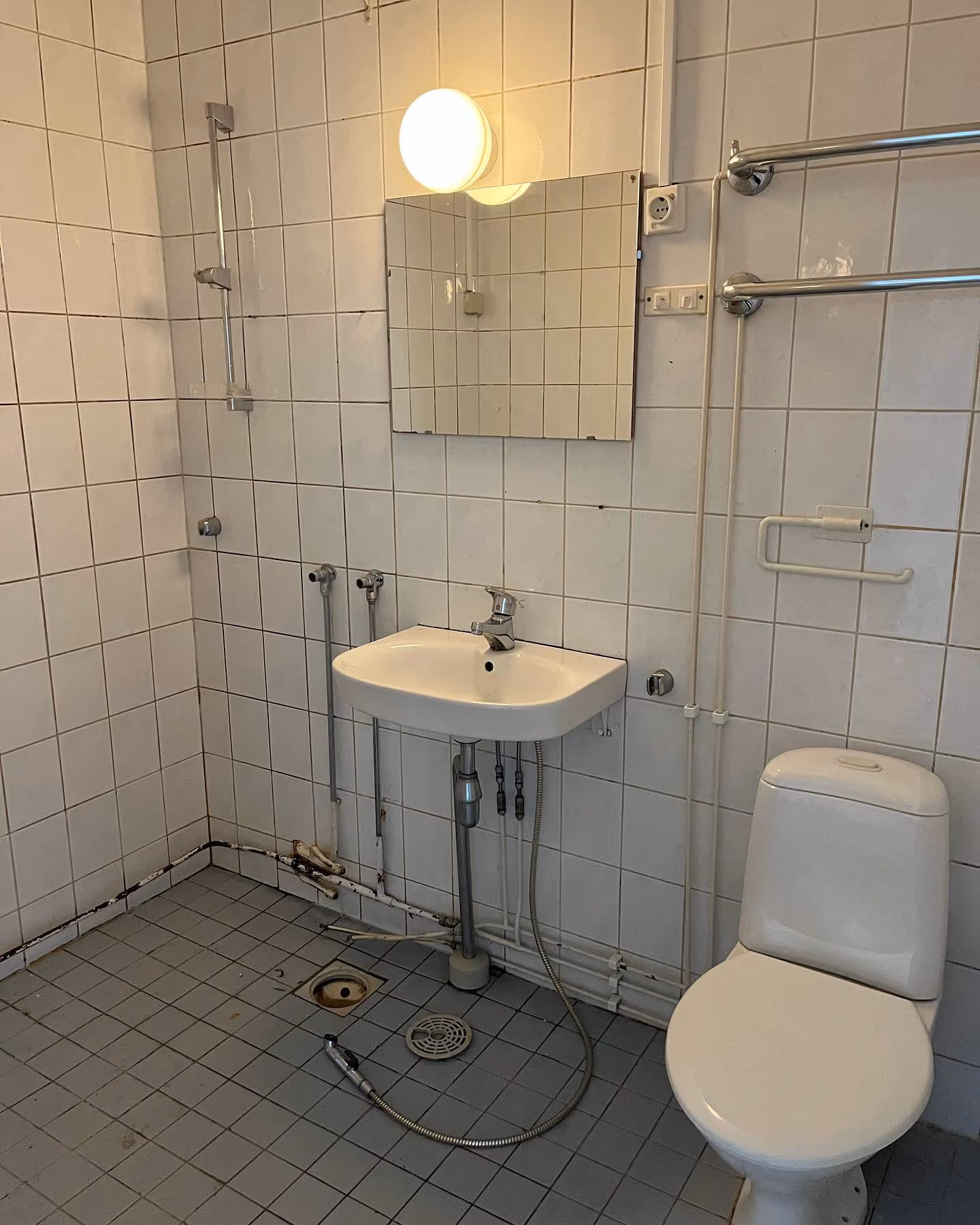 Pieni kylpyhuone, jossa on valkoiset kaakeliseinät, pesuallas, wc-istuin ja näkyvät putkistot.