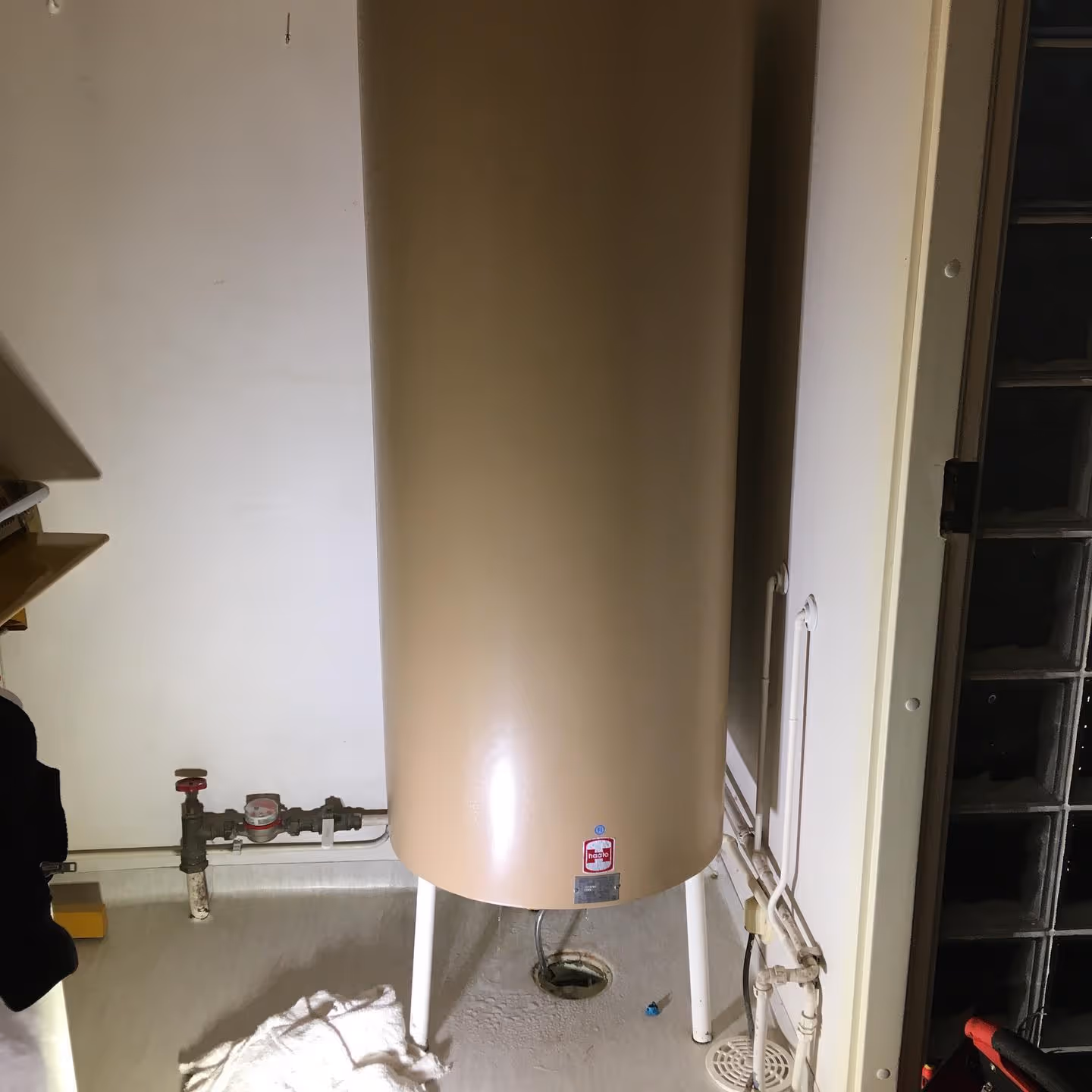 Beige lämminvesivaraaja seisoo teknisessä tilassa, jossa näkyy myös putkisto ja vesimittari.