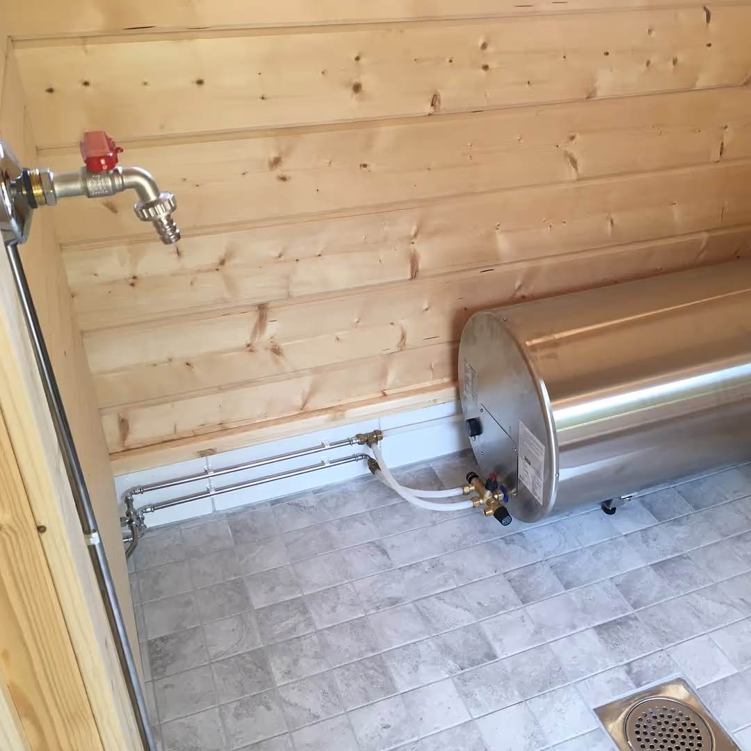 Puuseinäisessä huoneessa on laattalattia, vaakasuora lämminvesivaraaja ja seinään asennettu vesihana putkistoineen.