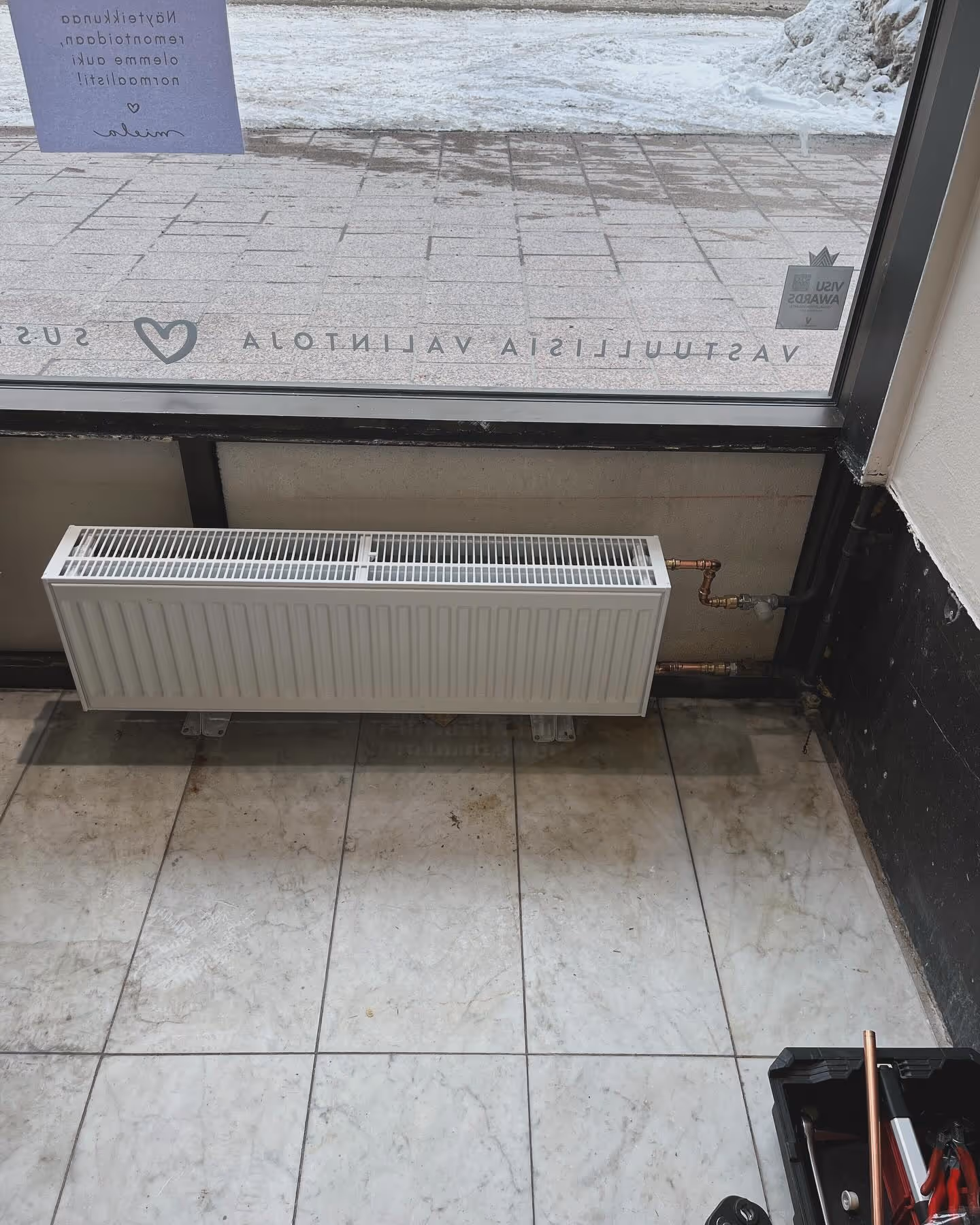 Valkoinen patteri on asennettu suuren ikkunan alle, ja siihen on liitetty kupariputket laatoitetulla lattialla.