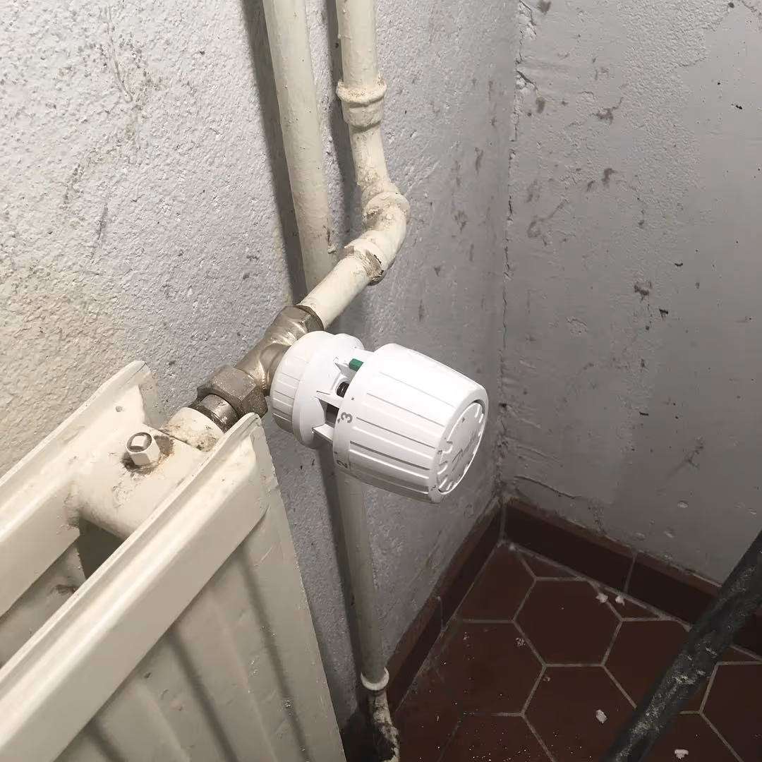 Valkoinen patteri ja termostaattiventtiili on liitetty putkiin harmaalla seinällä.