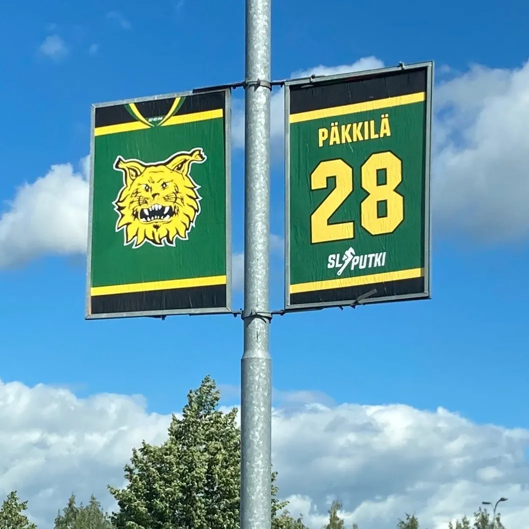 Kaksi vihreä-keltaista banneria roikkuu pylväässä, joista toisessa on keltainen ilveksen pää ja toisessa numero 28 sekä sana 'PUTKI'.