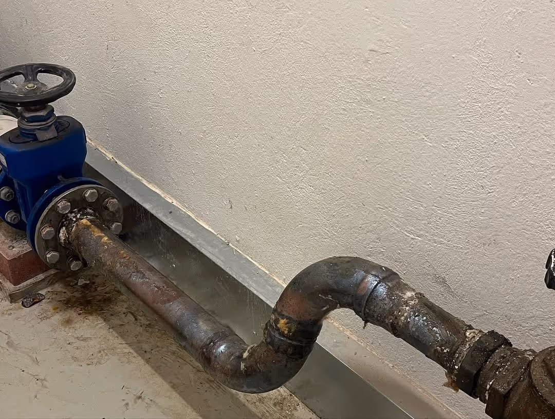 Ruosteinen metalliputki, jossa on sininen venttiili ja korroosiota, kulkee seinän vieressä.
