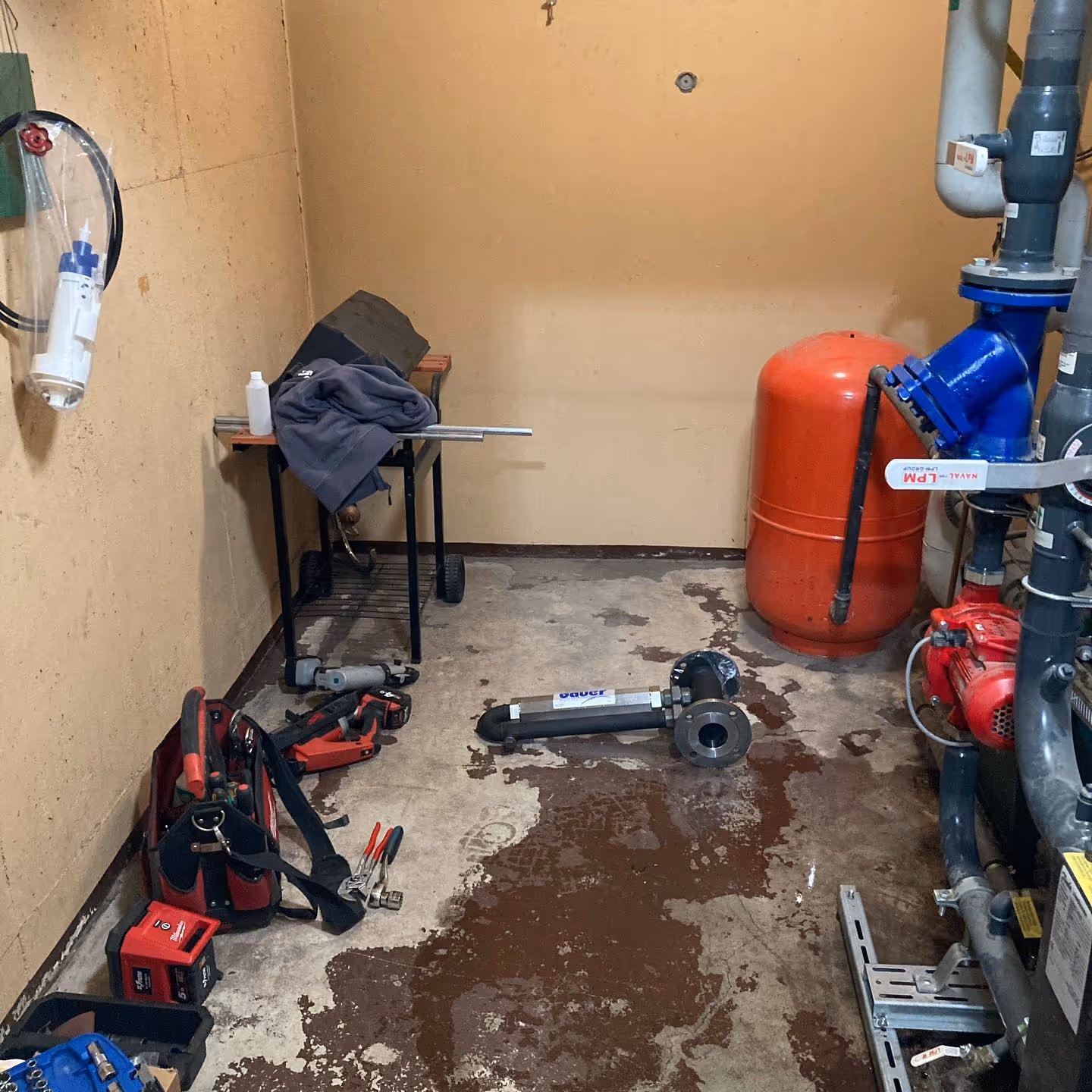 Huoneessa näkyy putkistoa, punainen painesäiliö, työkaluja ja lattialla ruskeaa nestettä.