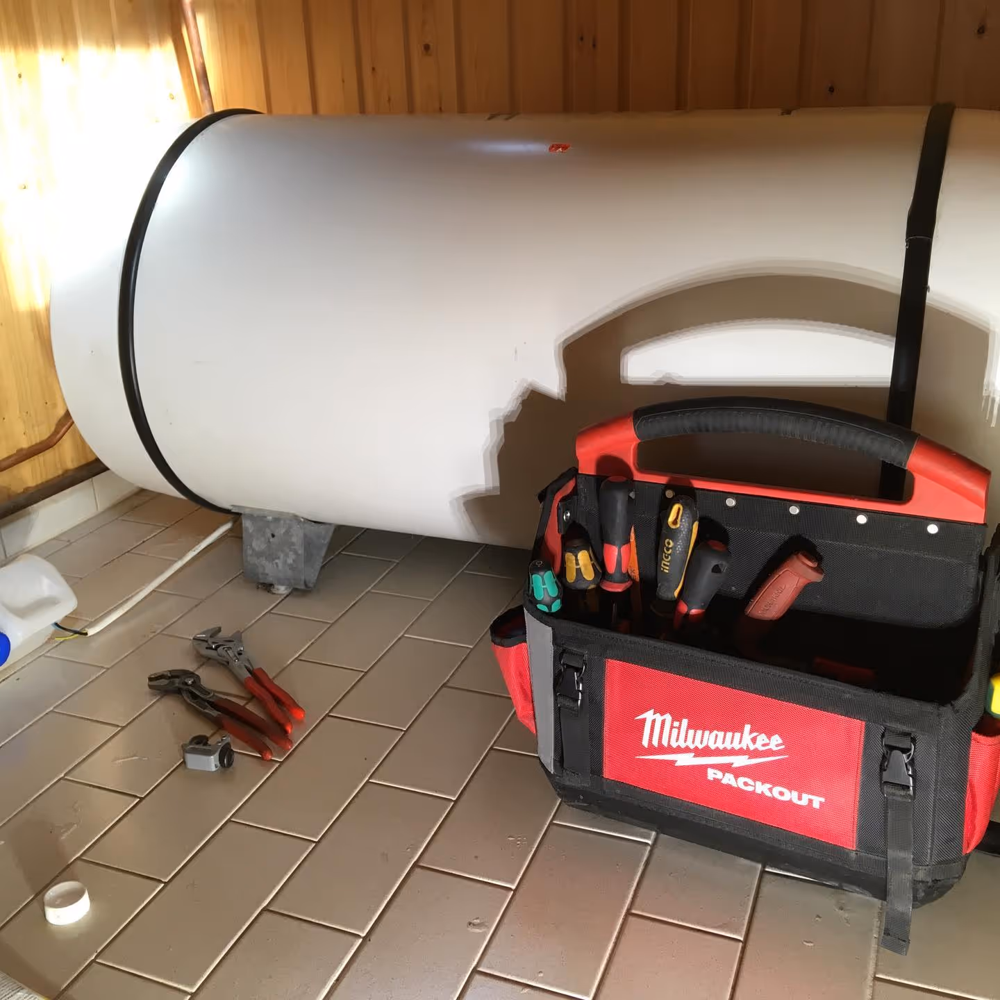 Valkoinen lämminvesivaraaja on asennettuna puuseinän edessä, ja sen vieressä on työkaluja putkitöitä varten.