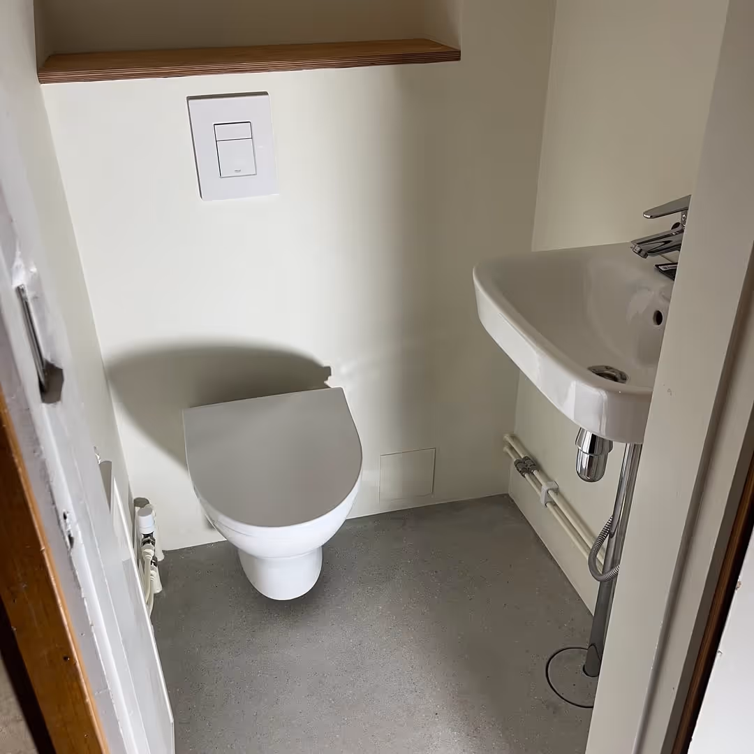 Pieni WC-tila, jossa on valkoinen WC-istuin, kulmapesuallas ja näkyvät putket.
